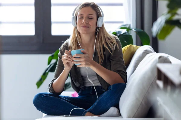 Mujer bastante joven escuchando música con auriculares mientras bebe una taza de café en el sofá en casa . — Foto de Stock