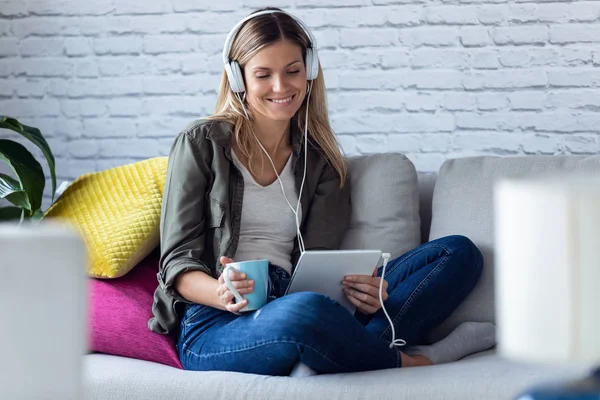 예쁜 아가씨가 디지탈 태블릿으로 음악을 듣고 집에 있는 소파에서 커피를 마시면서. — 스톡 사진