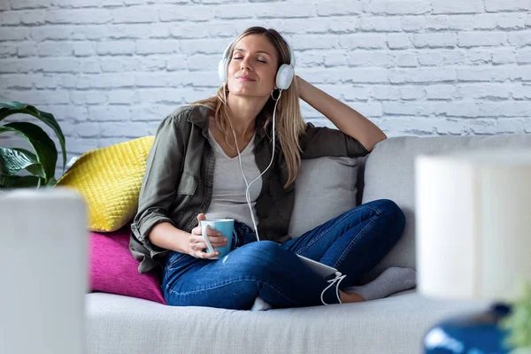 Mujer bastante joven escuchando música con auriculares mientras bebe una taza de café en el sofá en casa . — Foto de Stock