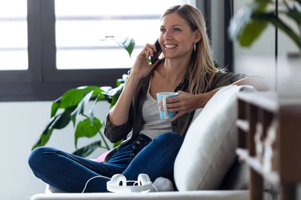 Mujer bastante joven hablando en el teléfono móvil mientras bebe una taza de café en el sofá en casa . — Foto de Stock