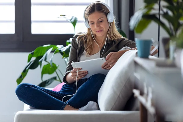 Mujer bastante joven escuchando música con auriculares y su tableta digital mientras está sentada en el sofá en casa . — Foto de Stock