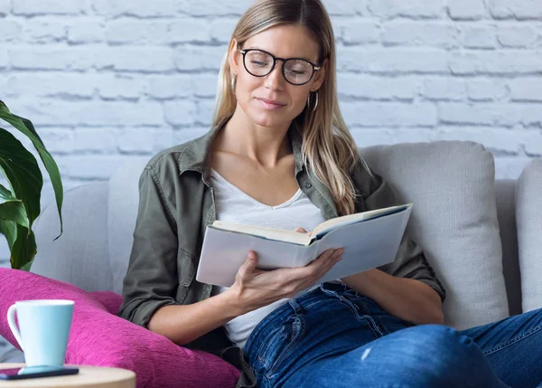 Söt ung kvinna läser en bok när hon sitter på soffan hemma. — Stockfoto