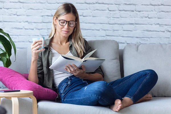 Mujer bastante joven leyendo un libro y bebiendo una taza de café mientras se sienta en el sofá en casa . — Foto de Stock