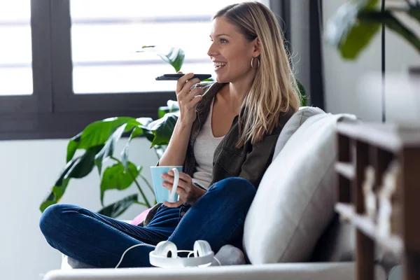 Mujer bastante joven hablando en el teléfono móvil con las manos libres mientras toma una taza de café en el sofá en casa . — Foto de Stock