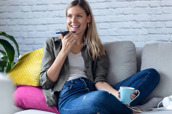 Mujer bastante joven hablando en el teléfono móvil con las manos libres mientras toma una taza de café en el sofá en casa . — Foto de Stock