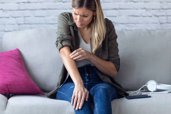 Uncomfortable young woman scratching her arm while sitting on the sofa at home. — Stock Photo, Image