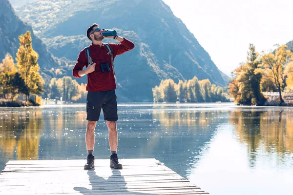 Vandrare ung man resenär med ryggsäck dricksvatten när du står framför sjön. — Stockfoto