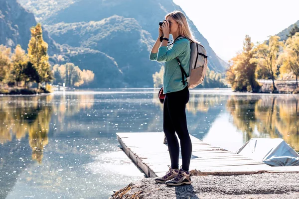 Muito jovem viajante com mochila tirando fotos com câmera no lago . — Fotografia de Stock