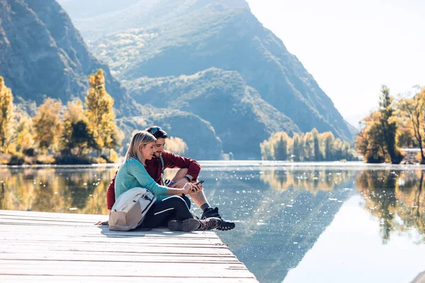 Två vandrare som använder mobiltelefon när de sitter framför sjön i fjällen. — Stockfoto