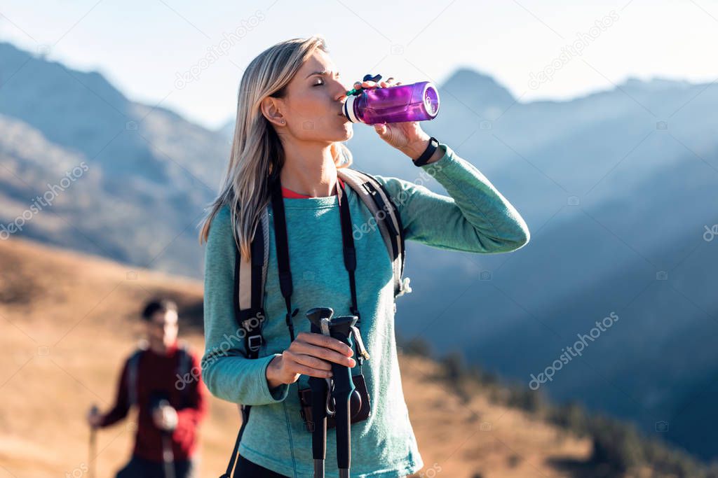 Pretty young woman traveler with backpack drinking water while standing on mountain.