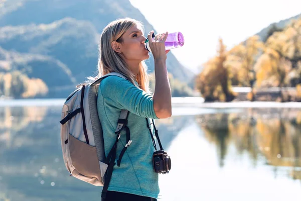 Muito jovem viajante com mochila bebendo água enquanto estava em frente ao lago . — Fotografia de Stock