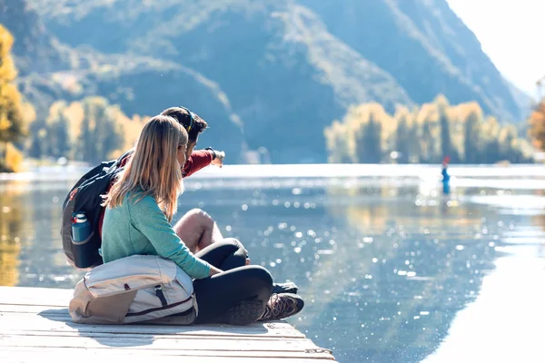 Due escursionisti di viaggio seduti mentre l'uomo indica qualcosa nel paesaggio del lago in montagna . — Foto Stock