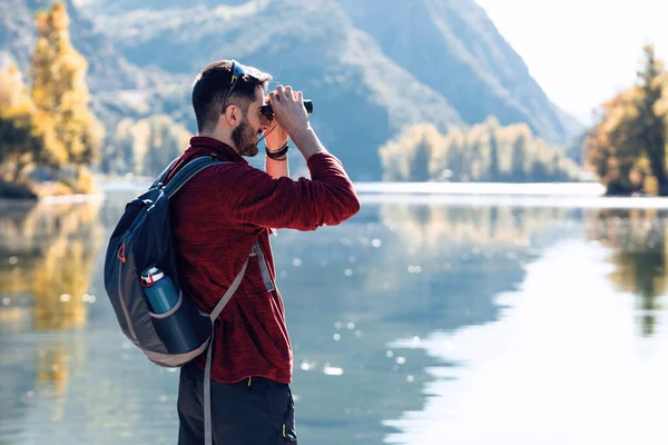 Vandrare ung man resenär med ryggsäck tittar landskapet med kikare i sjön. — Stockfoto