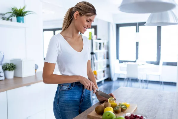 Mujer bastante joven cortando limones para preparar bebidas desintoxicantes en la cocina en casa . — Foto de Stock