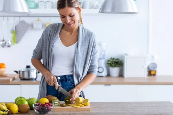 Mooie jonge vrouw snijden kiwi voor het bereiden van detox drank in de keuken thuis. — Stockfoto