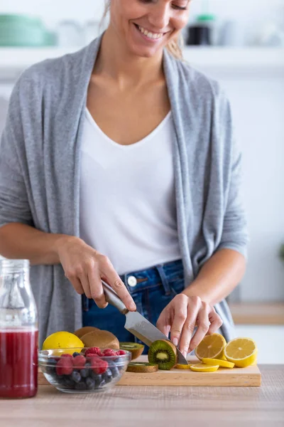 Mooie jonge vrouw snijden kiwi voor het bereiden van detox drank in de keuken thuis. — Stockfoto
