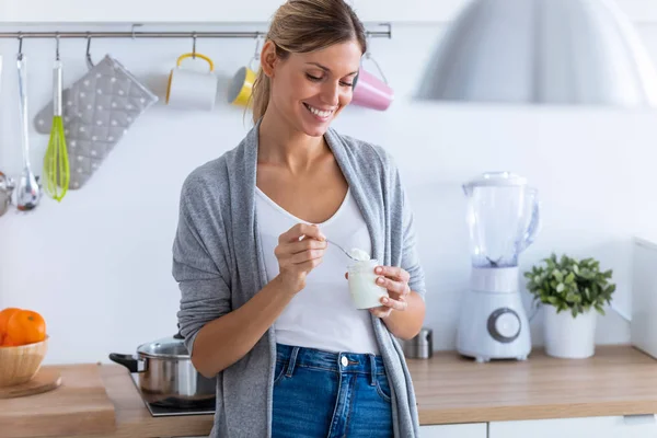 Wanita muda cantik makan yogurt saat berdiri di dapur di rumah . — Stok Foto