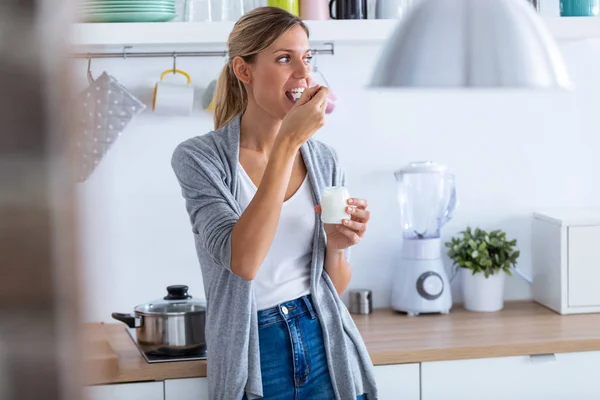 Wanita muda cantik makan yogurt saat berdiri di dapur di rumah . — Stok Foto