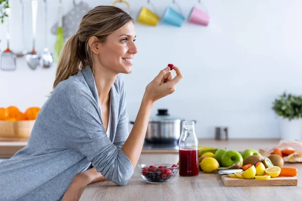 Hezká mladá žena jíst červené bobule, zatímco sedí v kuchyni doma. — Stock fotografie