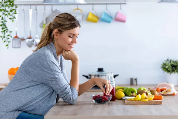 Mooie jonge vrouw die rode bessen eet terwijl ze thuis in de keuken zit. — Stockfoto