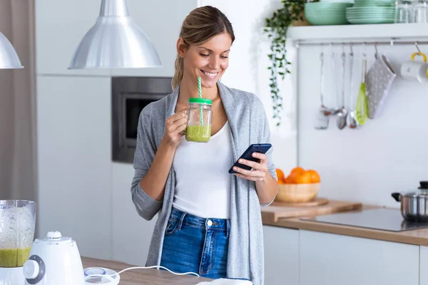 Mujer bastante joven usando su teléfono móvil mientras bebe jugo de desintoxicación en la cocina en casa . — Foto de Stock