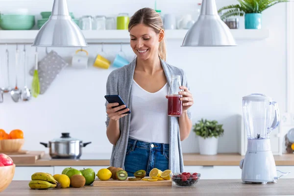 Mujer bastante joven usando su teléfono móvil mientras bebe jugo de frutas en la cocina en casa . —  Fotos de Stock