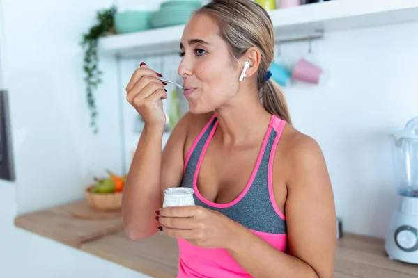 Pretty young woman eating yogurt while listening to music with wireless earphones in the kitchen at home. — Stock Photo, Image