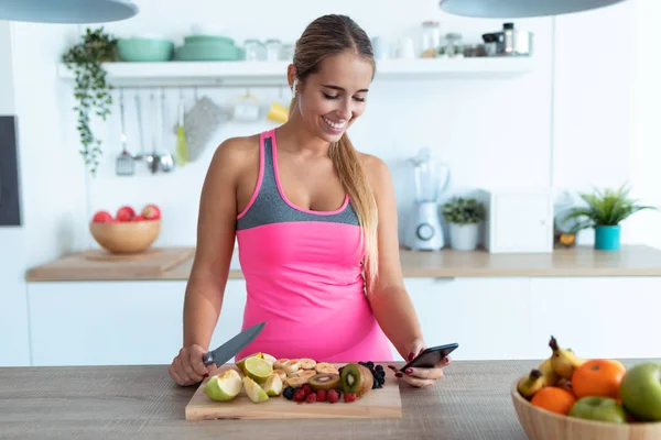 Mulher bonita usando seu telefone celular enquanto prepara suco de desintoxicação na cozinha em casa . — Fotografia de Stock