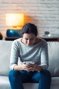 Oturma odasındaki kanepede otururken cep telefonunu kullanan gülümseyen genç bir kadın..