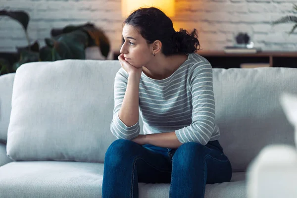 Tråkig ung kvinna tittar till sidan och tänker medan du sitter på soffan hemma. — Stockfoto