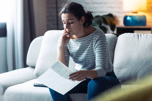 Orolig ung kvinna tittar på räkningar och tänker på sina problem när hon sitter på soffan hemma. — Stockfoto