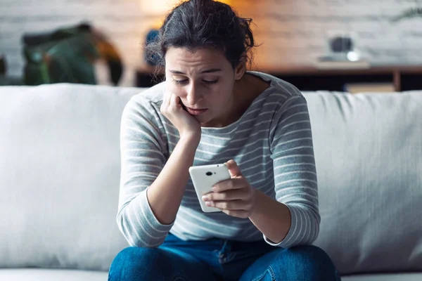 Jovem preocupada usando seu telefone celular enquanto se senta no sofá na sala de estar em casa . — Fotografia de Stock