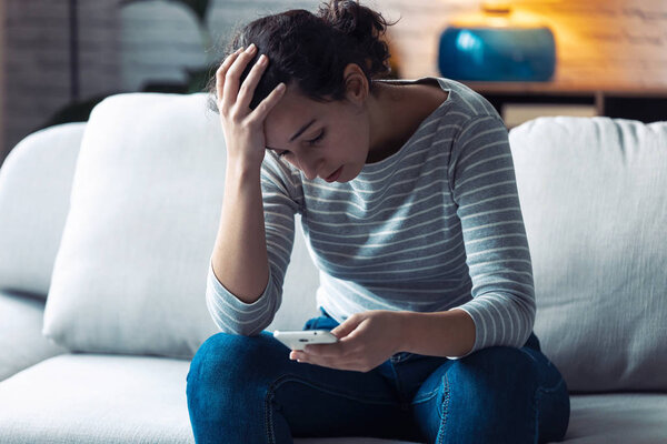 Worried young woman using her mobile phone while sitting on sofa in the living room at home.