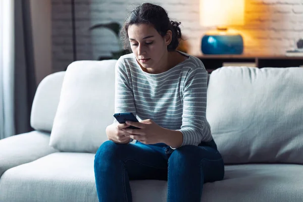 Jovem mulher séria usando seu telefone celular enquanto se senta no sofá na sala de estar em casa . — Fotografia de Stock
