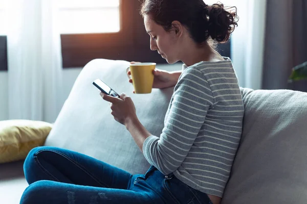 Серйозна молода жінка використовує свій мобільний телефон, сидячи на дивані у вітальні вдома . — стокове фото