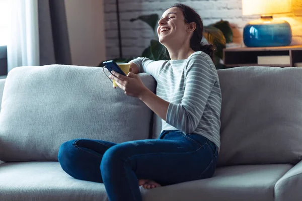 Sorrindo jovem mulher usando seu telefone celular enquanto sentado no sofá na sala de estar em casa . — Fotografia de Stock