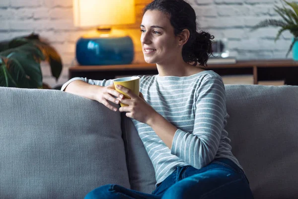 Lächelnde junge Frau, die zu Hause auf dem Sofa im Wohnzimmer sitzt und zur Seite schaut — Stockfoto