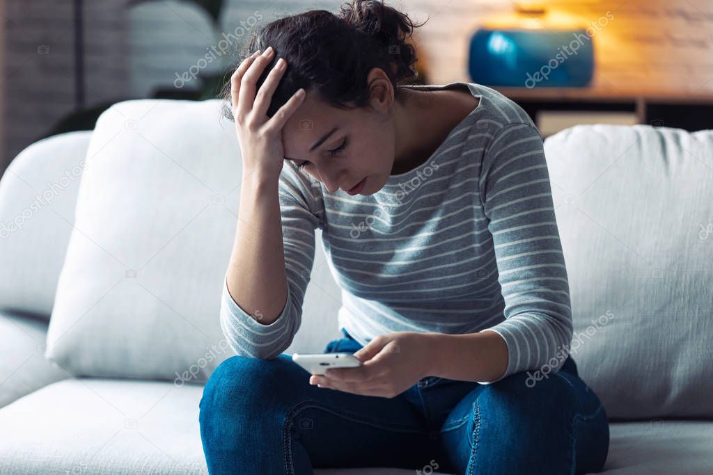 Worried young woman using her mobile phone while sitting on sofa in the living room at home.