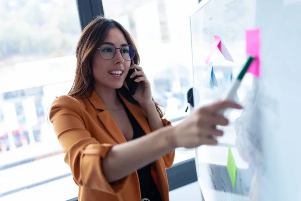 Business young woman talking on mobile phone while working on white board with post it stickers in the office. — Stock Photo, Image