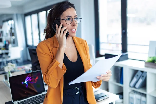 Affärs ung kvinna talar på mobiltelefon medan du håller papper på kontoret. — Stockfoto