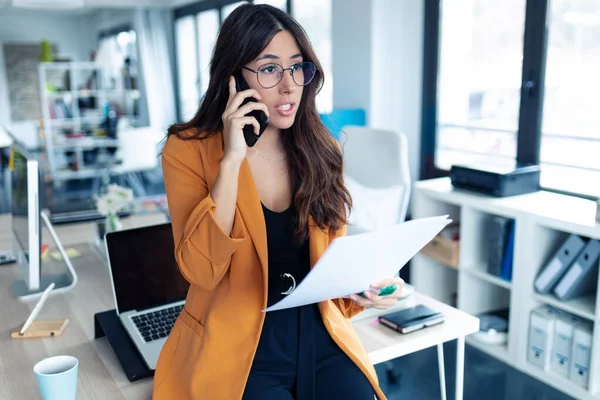 Üzleti fiatal nő beszél a mobiltelefon, miközben kezében papírok az irodában. — Stock Fotó