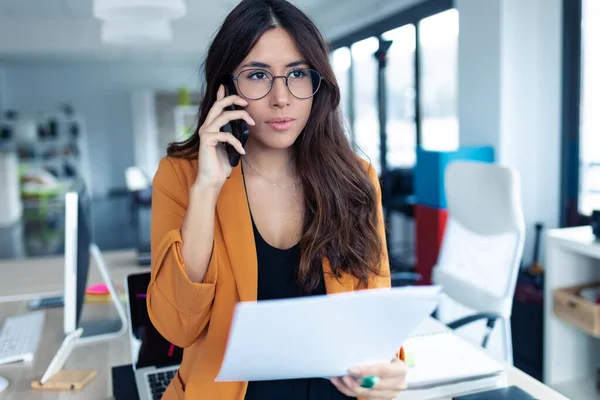Üzleti fiatal nő beszél a mobiltelefon, miközben kezében papírok az irodában. — Stock Fotó