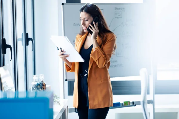 Femeie de afaceri care vorbește pe telefonul mobil în timp ce ține hârtii la birou . — Fotografie, imagine de stoc