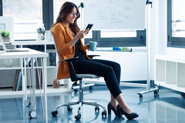 Femeie tânără de afaceri care își folosește telefonul mobil și bea cafea în timp ce stă la birou . — Fotografie, imagine de stoc