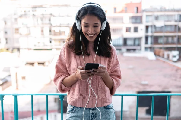 Mujer bastante joven escuchando música con teléfono inteligente mientras está de pie en la azotea en casa . — Foto de Stock