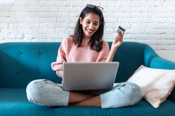Mooie jonge vrouw winkelen online met credit card en laptop tijdens het zitten op de bank thuis. — Stockfoto