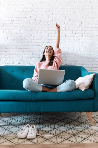 Mujer bastante joven celebrando algo mientras usa su computadora portátil en el sofá en casa . — Foto de Stock