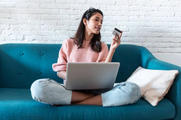 Mujer bastante joven de compras en línea con tarjeta de crédito y portátil mientras está sentado en el sofá en casa . — Foto de Stock