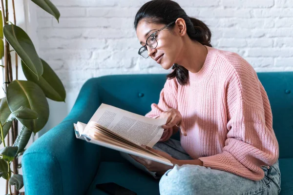 Söt ung kvinna läser en bok när hon sitter på soffan hemma. — Stockfoto