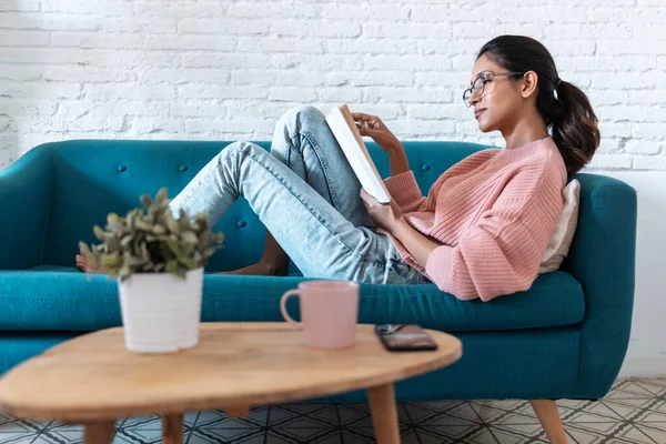 Bella giovane donna che legge un libro mentre è seduta sul divano a casa . — Foto Stock